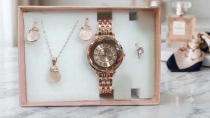 A rose gold jewelry gift set displayed inside a pink box on a white foam insert, featuring a watch, necklace, earrings, and ring on a marble surface.