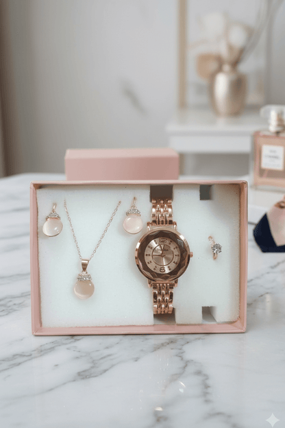 A rose gold jewelry gift set displayed inside a pink box on a white foam insert, featuring a watch, necklace, earrings, and ring on a marble surface.