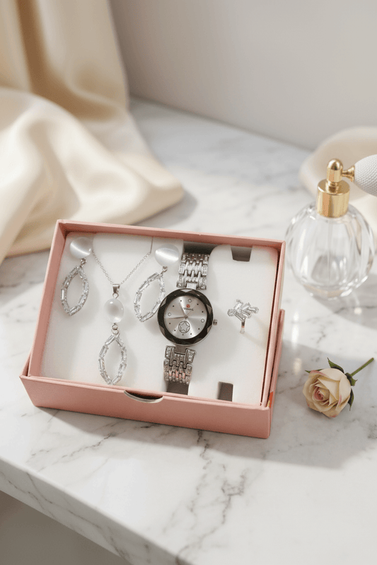 A jewelry and watch gift set displayed in an open blush pink box on a white marble surface, with draped cream fabric, a perfume bottle, and a small rose around it.
