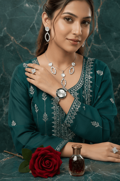 A woman wearing a silver-toned jewelry set including a necklace, earrings, ring, and watch, paired with a dark green embroidered kurta. She is posed on a white marble surface with draped cream fabric and a perfume bottle in the background.