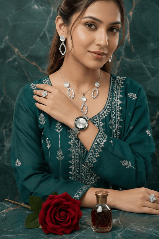 A woman wearing a silver-toned jewelry set including a necklace, earrings, ring, and watch, paired with a dark green embroidered kurta. She is posed on a white marble surface with draped cream fabric and a perfume bottle in the background.